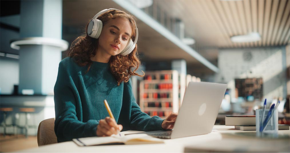 Girl wearing headphones writing in a notebook while studying at a desk with a laptop in a library